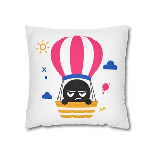 Könnte beinhalten: Ein quadratisches weißes Dekokissen mit einer Grafik einer schwarzen Figur mit mürrischem Gesichtsausdruck in einem Heißluftballon mit rosa-weißen Streifen. Das Design enthält eine gelbe Sonne, blaue Wolken und kleine geometrische Formen in Blau und Rosa.