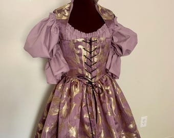 May include: A lavender and gold Renaissance-style gown. The bodice features a black lace-up front and gold floral detailing. The skirt is a full length with a matching gold floral pattern. The sleeves are puffed lavender fabric.