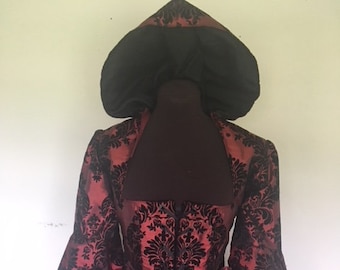 May include: A long, red and black brocade dress with a black velvet hood. The dress has a fitted bodice and a full skirt. The hood is attached to the dress at the neckline.