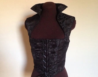 May include: A black sleeveless corset with a lace-up front and a high, pointed collar. The corset is made of a brocade fabric with a floral pattern.