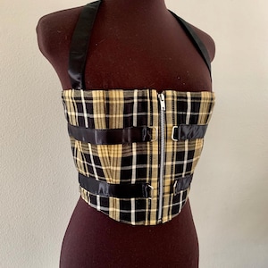 May include: A plaid corset top in black, white, and gold, displayed on a mannequin. The top features a front zipper, black straps, and buckle details. The halter straps are black and shiny.