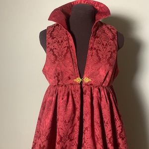May include: A red damask fabric costume with a high collar and a gold clasp. The costume is displayed on a mannequin. The fabric has a detailed floral pattern. The costume appears to be a historical or theatrical garment.