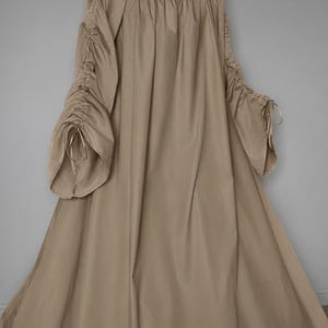 May include: A floor-length beige dress featuring a gathered scoop neckline and long, loose sleeves. The sleeves are detailed with vertical drawstring channels that create a ruched effect, finished with thin ties at the flared cuffs.