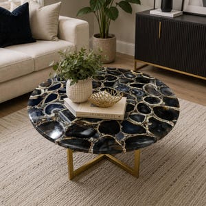 May include: A round coffee table with a polished, dark blue and black stone top. The table has a gold-colored metal base and sits on a light beige rug. A white sofa and black cabinet are in the background.