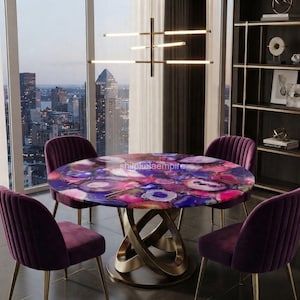 Op de afbeelding: Een ronde eettafel met een veelkleurig agaatstenen blad in tinten paars, roze en blauw, ondersteund door een goudkleurig metalen onderstel met in elkaar grijpende gebogen poten, omringd door vier paarse fluwelen stoelen met verticale stiksels.