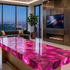 May include: A rectangular dining table featuring a polished surface made of vibrant pink agate slices with natural banding patterns. The table has a solid base and is positioned in a room with a city view. The text shilpkalaempire is visible.