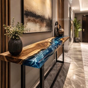 Puede incluir: Una consola larga de madera con un centro de resina epoxi azul en forma de río. La mesa tiene una estructura de metal oscuro y está colocada en un pasillo. Un jarrón negro con flores está sobre la mesa. El texto "shipkalaempire" es visible.