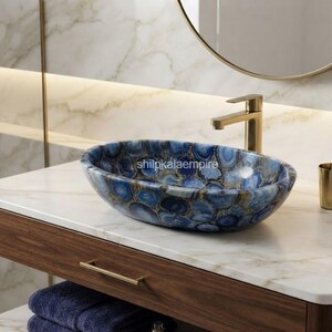May include: An oval-shaped bathroom sink crafted from blue agate stone with natural banding patterns. The basin features a polished finish and is centered on a white marble countertop. The text shilpkalaempire is visible on the basin surface.