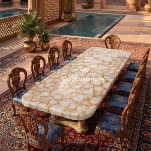 May include: A rectangular dining table with a white stone top featuring gold veining, supported by a pedestal base. Ten carved wooden chairs with blue patterned upholstery are arranged around the table. The text shilpkalaempire is visible.