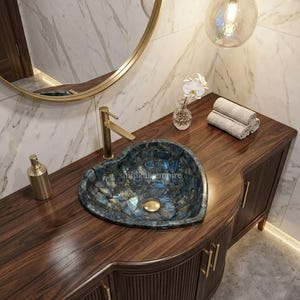 Op de afbeelding: Een hartvormige blauwe stenen waskom met een gouden kraan en afvoer, geplaatst op een donkerhouten wastafelmeubel met verticale latten en gouden handgrepen. Het meubel heeft een glad houten oppervlak, een ronde spiegel en een glazen hanglamp.