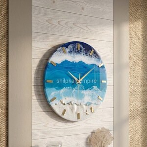 May include: A round wall clock featuring a resin ocean scene with shades of deep blue, turquoise, and white foam waves. The bottom section includes sand and small seashells. It has gold-coloured hands and markers, with the text shilpkaempire visible.