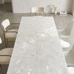 May include: A rectangular dining table featuring a white mother-of-pearl inlay surface arranged in a sunburst pattern with a central circular motif. The text shilpkalaempire is printed in the center of the table surface. The table has a flat top.