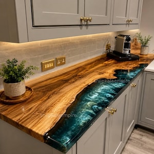 May include: A custom kitchen countertop with a live edge design. The countertop features a rich, natural wood grain on one side and a deep teal epoxy resin river on the other. The cabinets are gray with gold hardware.
