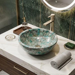 May include: A round vessel sink crafted from polished green and white stone fragments, featuring a smooth interior basin and a central drain, positioned on a white marble countertop with a gold faucet and a rolled grey towel nearby.