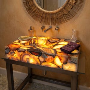 May include: A backlit agate stone sink with a warm, amber glow. The sink is supported by a dark metal frame with a glass shelf. A decorative mirror and fixtures are visible in the background. A small bottle of perfume, soap, and a towel are on the sink.