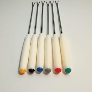 May include: A set of six metal fondue forks with long, slender silver-toned shafts and white cylindrical handles. Each handle features a distinctively colored end cap in yellow, black, blue, gray, red, and green for easy identification during use.