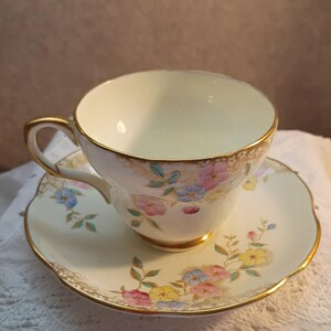 May include: Cream-colored teacup and saucer set with floral designs in pink, blue, and yellow. The cup and saucer have gold trim. The saucer has a scalloped edge. The teacup has a curved handle.