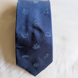 May include: A dark blue necktie featuring a fine ribbed texture and a repeating pattern of light blue embroidered animal icons, including birds, fish, and mythical creatures scattered across the fabric surface in a structured layout.