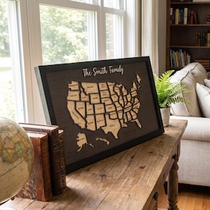 May include: A framed wooden wall art piece featuring a laser-cut map of the United States with individual state outlines and labels. The text The Smith Family is displayed in white script at the top against a dark brown textured background.