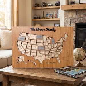 May include: A rectangular wooden USA travel map featuring a laser-cut design with the text The Brown Family at the top and USA TRAVEL MAP at the bottom. The map displays individual state outlines with some states highlighted in a grey finish.