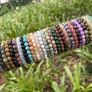 Può includere: Una collezione di numerosi braccialetti con perline rotonde di pietre preziose disposti in fila su un espositore cilindrico. I braccialetti presentano una varietà di colori e consistenze, tra cui tonalità di verde, marrone, nero, bianco, rosa, viola e blu.