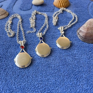 May include: Three silver-toned circular locket pendants on matching silver-toned chains. The left locket features a small red stone at the top, the center locket has a small blue stone, and the right locket is plain silver-toned metal without a stone.
