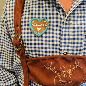 May include: A blue and white gingham button-down shirt worn with brown suede suspenders and a brown suede chest panel featuring an embroidered stag. A heart-shaped gingerbread cookie with blue icing and the text WIESN 26 is pinned to the shirt.