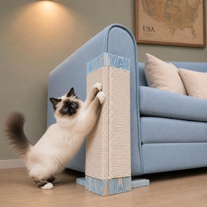 Puede incluir: Un gato de color crema y marrón oscuro con ojos azules se apoya en sus patas traseras mientras araña un rascador vertical rectangular. El poste tiene tapas de madera teñidas de azul claro en los extremos y un centro envuelto en cuerda de sisal.