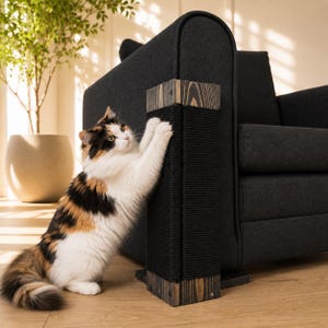Puede incluir: Un rascador vertical para gatos con marco de madera oscura y cuerda negra, colocado contra la esquina de un sofá tapizado de color gris oscuro. El poste tiene un diseño rectangular con vetas de madera visibles y herrajes metálicos.