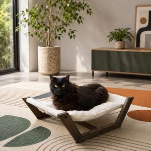 May include: A fluffy black cat with yellow eyes resting on a white fabric pet bed. The bed features a dark brown wooden frame with a V-shaped base design. The cat is positioned in the center of the white cushion, which is suspended between the wooden supports.