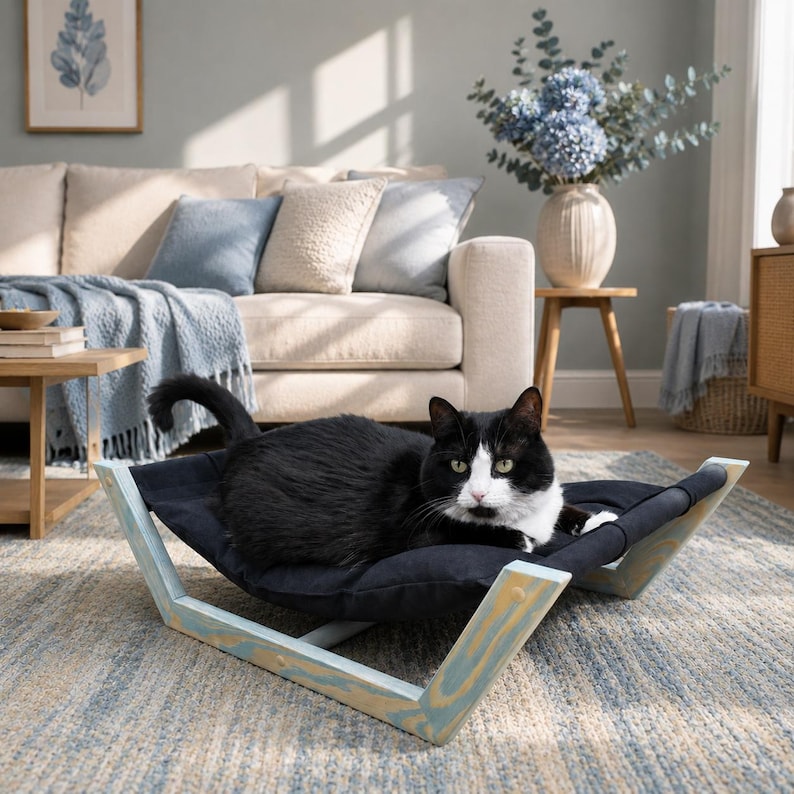 May include: A black and white cat resting on a black fabric hammock bed with a light blue wooden frame. The hammock features a curved design that suspends the fabric between two angled wooden supports, providing a raised surface for the cat.