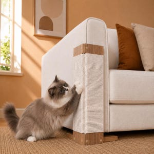 Puede incluir: Un rascador para gatos fijado a la esquina de un sofá de color claro. El rascador presenta una sección vertical de cuerda de sisal blanca enrollada alrededor de una columna central, enmarcada por paneles de madera marrón claro en los extremos.