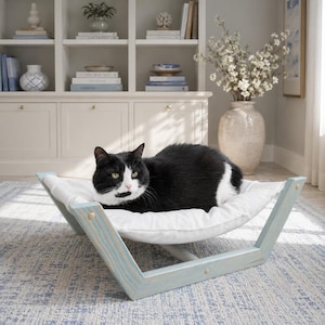 May include: A black and white cat resting on a pet hammock. The hammock features a light blue wooden frame with a distressed finish and a soft, white fabric bed suspended between the two angled side supports, creating a comfortable lounge area.