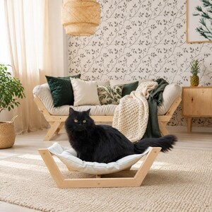 May include: A fluffy black cat sits in a light-colored wooden pet hammock with a soft white fabric bed. The hammock has a low-profile, open-frame design made of natural wood, providing a comfortable resting spot for a pet on the floor.