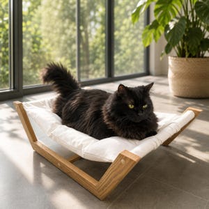 May include: A long-haired black cat resting on a white fabric hammock bed with a light brown wooden frame. The cat has green eyes and a fluffy tail. The bed is elevated on a sturdy wooden base, providing a comfortable spot for the feline to lounge.