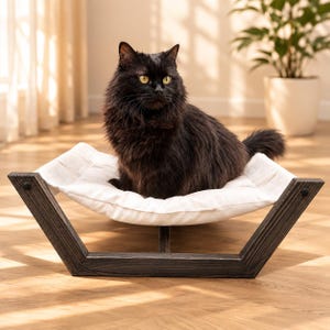 May include: A fluffy black cat with yellow eyes sitting on a white fabric hammock bed. The bed features a dark brown, weathered wooden frame with a V-shaped base design that supports the suspended, soft white cushion for resting.