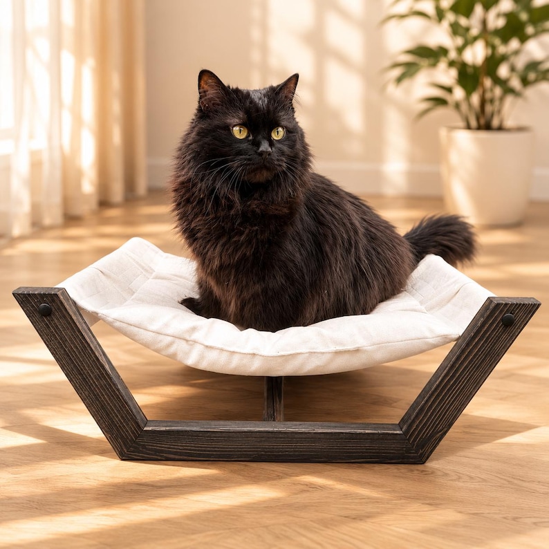 May include: A fluffy black cat with yellow eyes sitting on a white fabric hammock bed. The bed features a dark brown, weathered wooden frame with a V-shaped base design that supports the suspended, soft white cushion for resting.