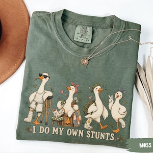 May include: A moss green short-sleeve t-shirt featuring a graphic of four white geese with various injuries and accessories. The text below the geese reads, I DO MY OWN STUNTS. The shirt has a crew neckline and a Comfort Colors brand label inside.