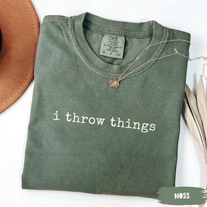 May include: A moss green short-sleeve t-shirt with a crew neckline and the text "i throw things" printed in white across the chest. The shirt features a Comfort Colors brand label at the inner neck and is paired with a delicate gold-tone necklace.