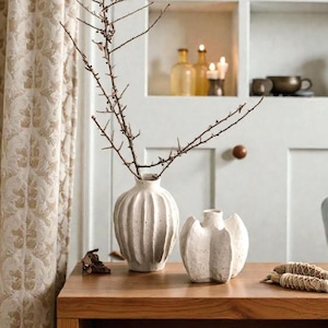 Könnte beinhalten: Ein Stillleben mit zwei weißen Keramikvasen auf einem Holztisch. Eine Vase ist hoch und gerippt und hält Zweige. Die andere ist eine kleinere, dekorative Vase. Ein neutralfarbener Vorhang und ein Einbauregal im Hintergrund.
