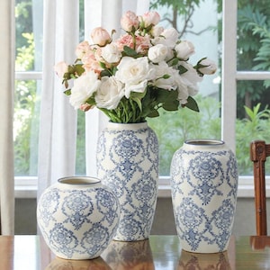 Puede incluir: Tres jarrones de cerámica blanca con un estampado floral azul. Los jarrones son de diferentes alturas y formas, con un ramo de rosas blancas y rosas en el jarrón más alto. Los jarrones están sobre una mesa de madera frente a una ventana.