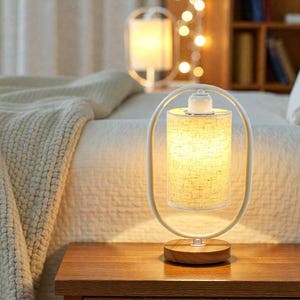 Puede incluir: Una lámpara de mesa con una pantalla cilíndrica de tela beige suspendida dentro de un marco ovalado de metal blanco. La lámpara está montada sobre una base circular de madera y emite una luz cálida desde el centro del marco.