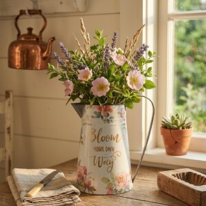 May include: A white metal pitcher with a floral design and the text "Bloom Your Own Way" holds a bouquet of pink flowers, lavender, and greenery. A copper kettle hangs in the background, and a small succulent sits in a terracotta pot.