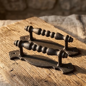 May include: Two dark, textured metal door handles with a spindle-shaped grip and a decorative, elongated backplate featuring scalloped edges and screw holes at each end. The handles have a matte, dark gray finish and a distinct, bumpy surface texture.