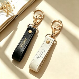 May include: Two rectangular keychains with gold-toned metal hardware, including a lobster clasp and a split ring. One keychain is black with the name Fiona embossed in gold, and the other is white with the name Nancy embossed in gold.