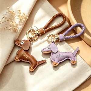 May include: Two dachshund-shaped keychains with braided loops. One is brown with the name Kian engraved, and the other is light purple with the name Alia engraved. Both feature gold-tone metal rings and hardware, with stitched edges and small white eye details.