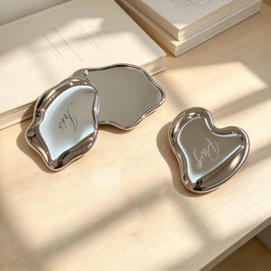 Könnte beinhalten: Drei silberfarbene, unregelmäßig geformte Metallschalen in flüssiger Optik mit reflektierenden Oberflächen. Zwei Schalen tragen eingravierte Namen, eine mit der Aufschrift Iris und die andere mit Paige, in glatter, polierter Ausführung.