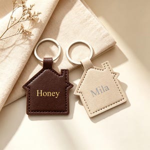 Può includere: Due portachiavi a forma di casa con anelli in metallo argentato. Uno è marrone scuro con il nome Honey impresso in oro, l'altro è color crema con il nome Mila impresso in grigio chiaro. Entrambi presentano cuciture visibili.