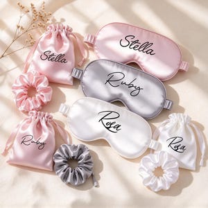May include: Three satin sleep masks, three drawstring pouches, and three scrunchies in pink, silver, and white. The items are personalized with the names Stella, Ruby, and Rosa in black script text printed on each piece.