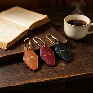 May include: Three diamond-shaped leather keychains with gold-toned metal hardware. The tan keychain is engraved with the name Stella, the maroon keychain with Katie, and the black keychain with Patty. Each features a gold-toned metal loop.